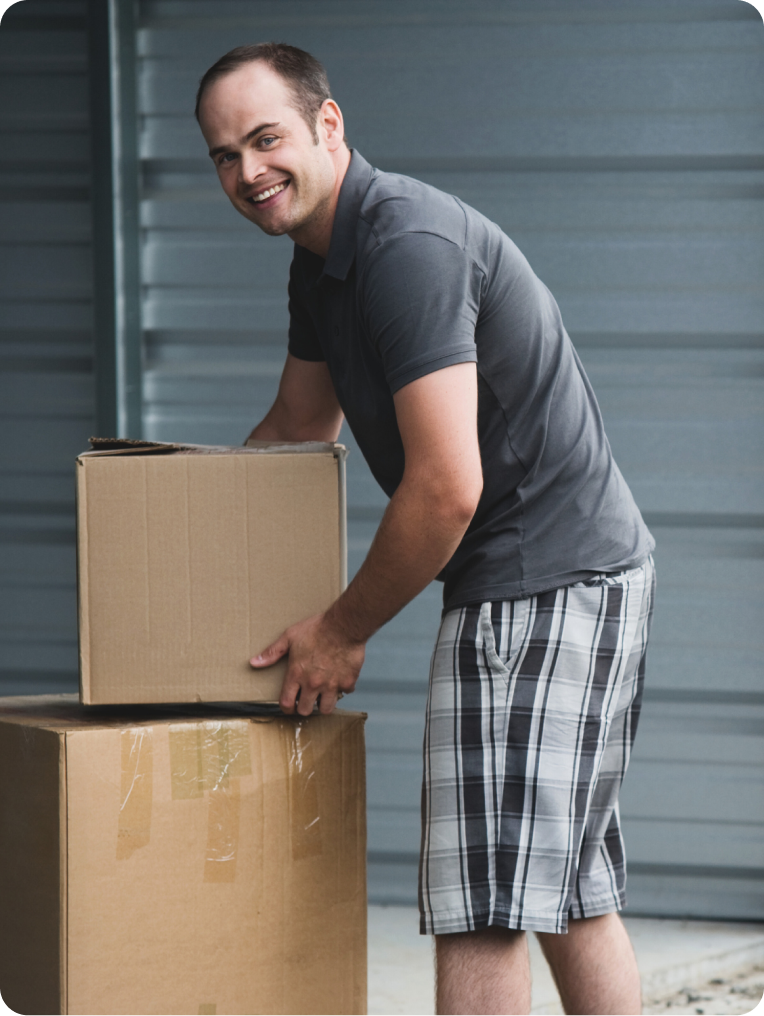 Smiling man in a gray shirt and plaid shorts lifting a cardboard box at Springfield Storage facility.